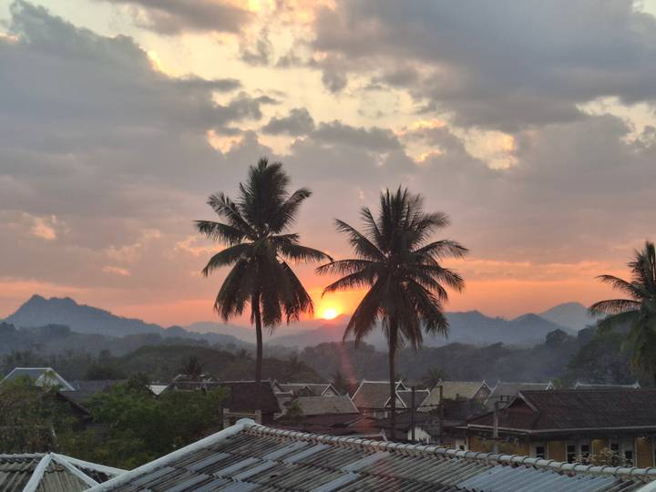 Silhouetted palm trees framing an orange sunset over misty mountains and rooftops.