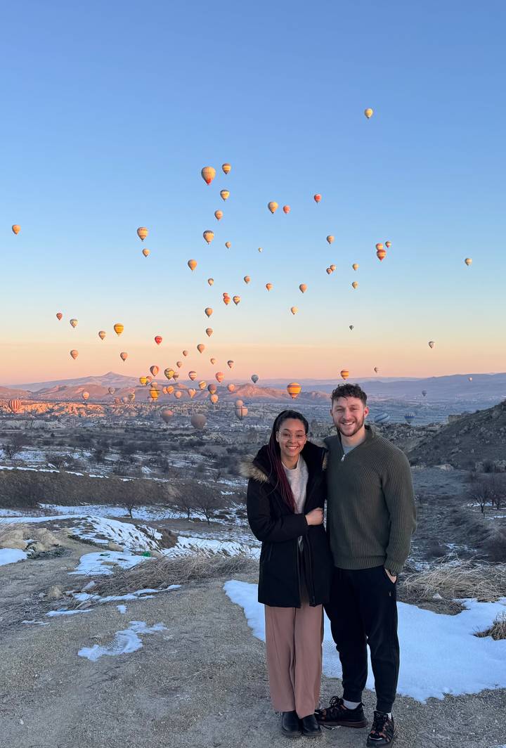 Couple smiling with dozens of colorful hot-air balloons rising over Cappadocia at sunrise.