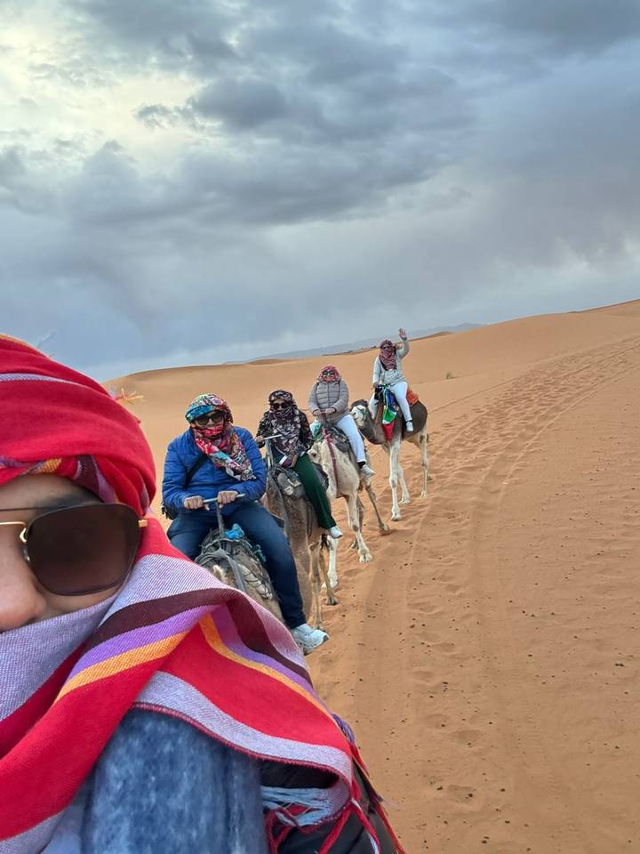 A line of tourists riding camels across rolling orange dunes, with the lead rider snapping a colourful scarf-covered selfie.