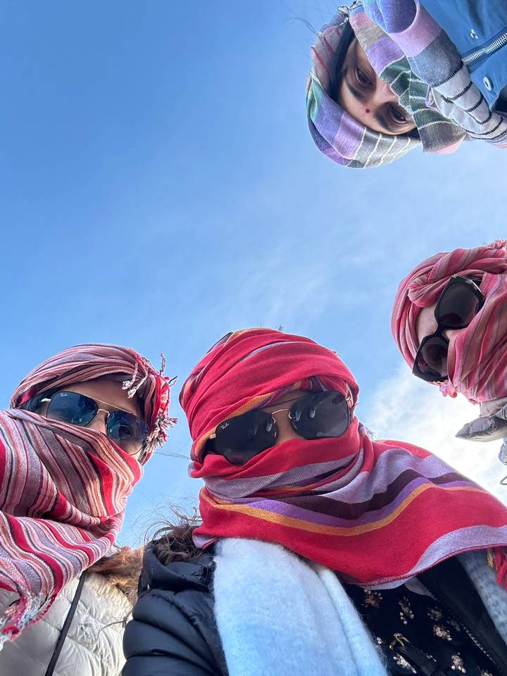 Three travellers in bright desert head-scarves and sunglasses smile up toward the camera against a blue sky.