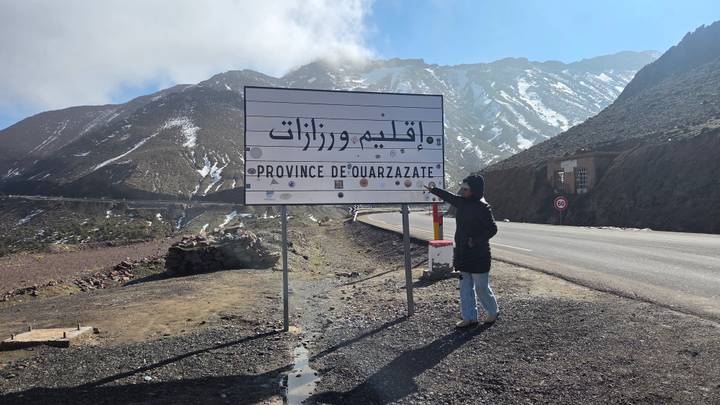 A traveller points at a bilingual road sign for the Province of Ouarzazate with snow-dusted peaks behind.