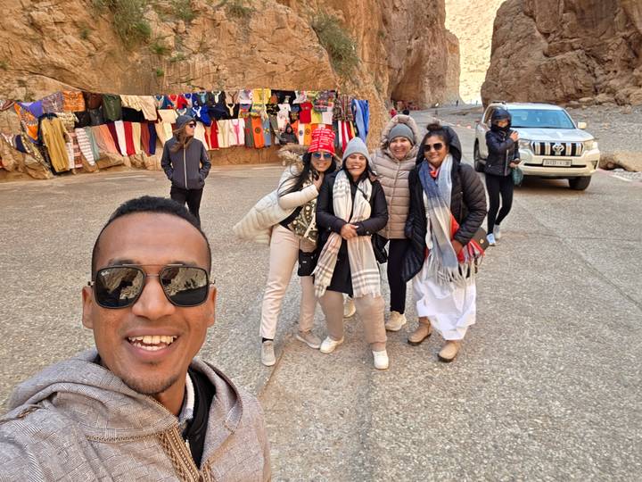 Smiling travellers snap a selfie beside market stalls of bright fabrics inside the rocky corridor of Todra Gorge.