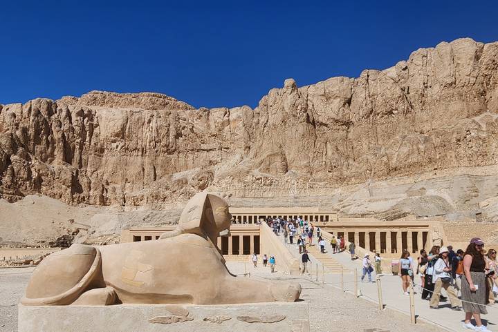 Wide view of the Mortuary Temple of Hatshepsut with tourists ascending the grand ramp, watched by a sphinx statue.