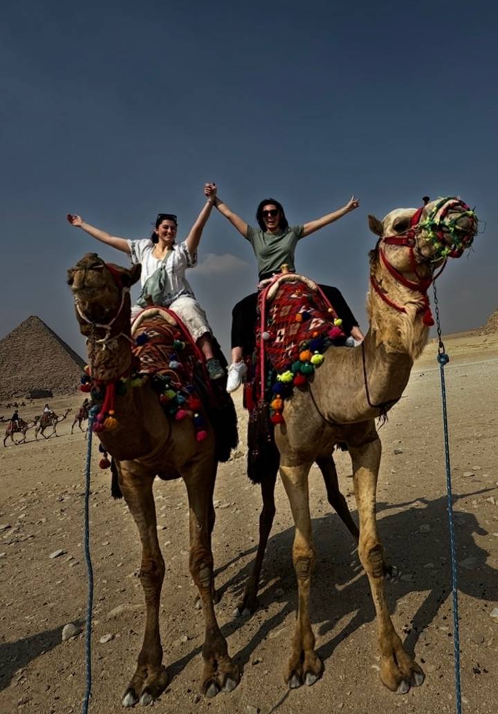Two joyful travelers pose atop decorated camels in the desert with the Pyramids of Giza visible behind them.