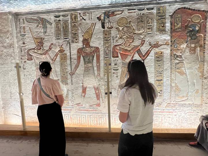 Visitors study vividly painted hieroglyphic walls inside an ancient Egyptian tomb or temple hall.