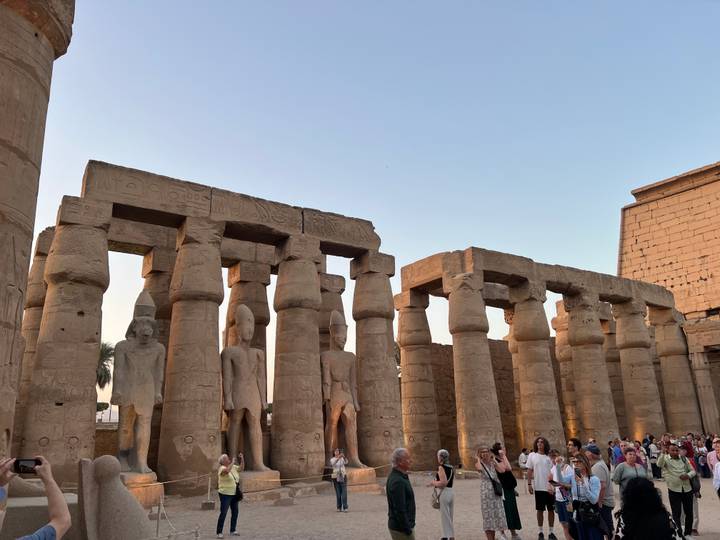Crowds gather among colossal statues and sandstone columns at Luxor Temple in the soft evening light.
