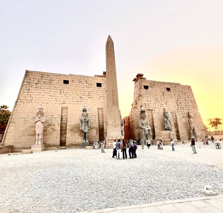 Obelisk and massive pylons of Luxor Temple glow against a colorful sunset sky while visitors explore the forecourt.