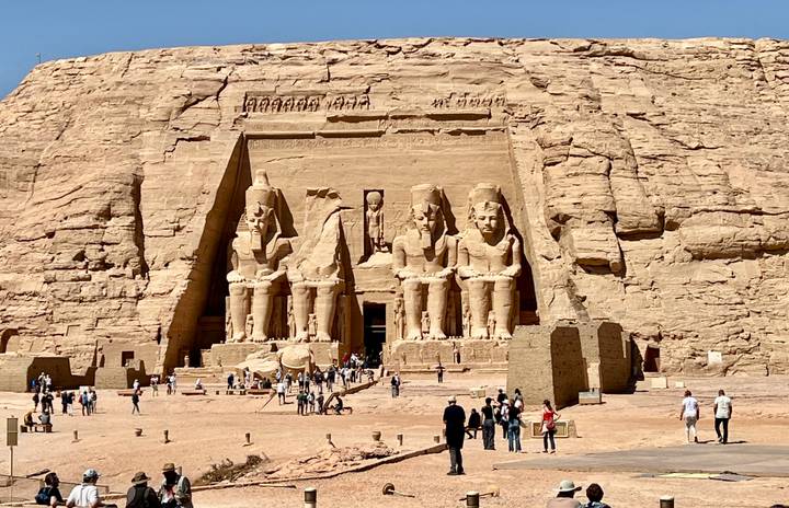 Grand rock-cut façade of Abu Simbel with four colossal seated statues overlooking a sandy forecourt filled with tourists.