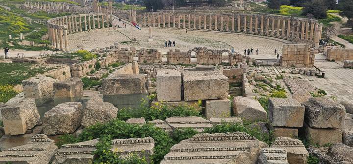 Panoramic view of Jerash’s oval forum ringed by standing columns with scattered visitors exploring the ancient site.