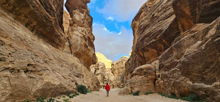 Narrow sandstone canyon in Petra with dramatic towering walls and a hiker in a red jacket walking the sandy floor.