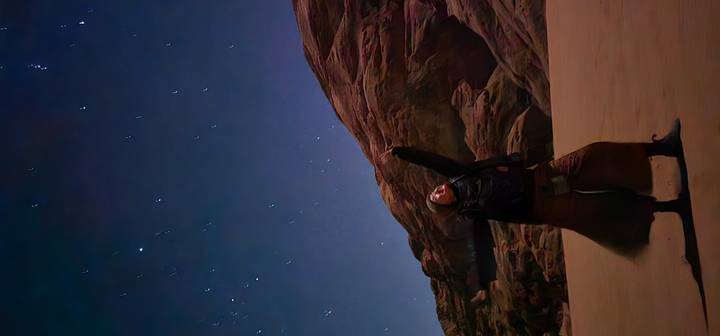 Nighttime desert scene with a traveler pointing toward a star-filled sky above rugged sandstone outcrops.