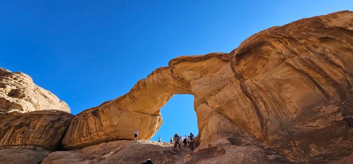 Spectacular sandstone arch in Wadi Rum with adventurers standing triumphantly on top against a vivid blue sky.