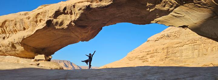 Person striking a joyful pose beneath a massive natural sandstone arch in Wadi Rum.