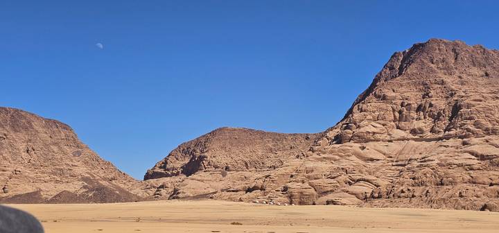 Wide desert valley framed by rugged sandstone mountains under a cloudless sky with the moon visible in daylight.