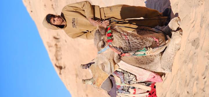 Bedouin camel handler in tan clothing standing beside a resting camel adorned with colorful reins in Wadi Rum.