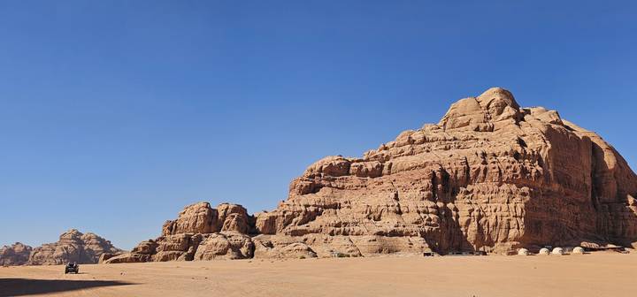 Expansive desert landscape with towering red sandstone massif under a clear blue sky and faint vehicle tracks in the sand.