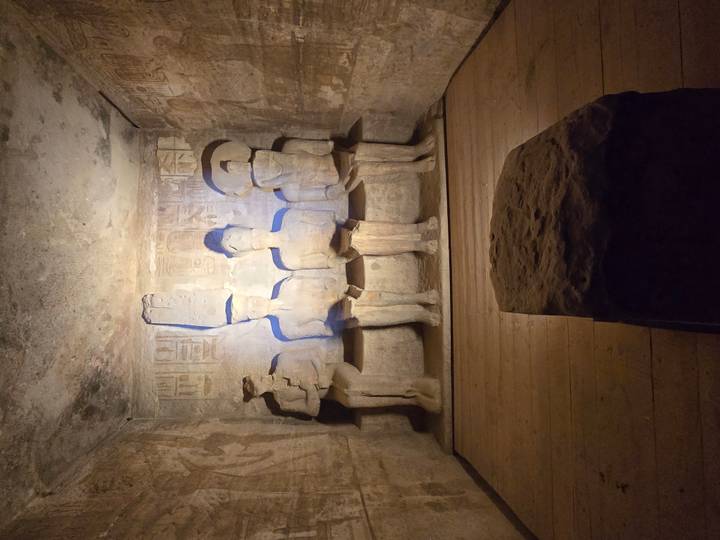 Dimly lit chamber of Abu Simbel with four seated statues carved into the sandstone wall, illuminated by spotlight.