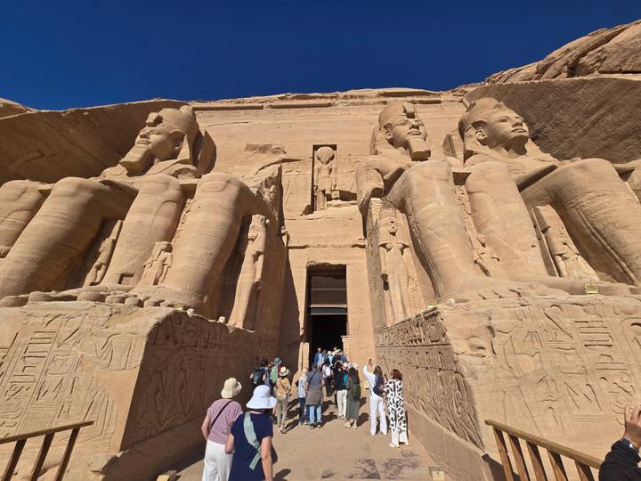 Grand façade of Abu Simbel temple with colossal Ramses II statues as tourists walk up the central stairway under clear blue sky.