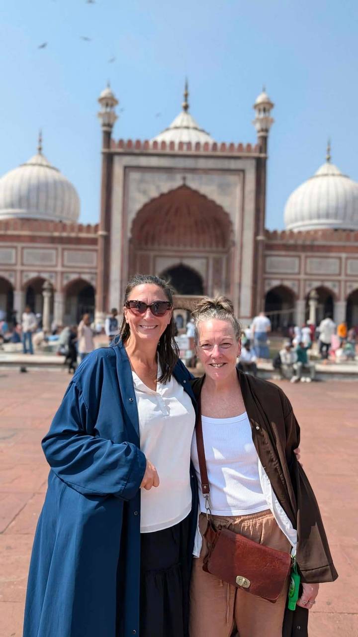 Two smiling women posing in front of a grand red-sandstone mosque courtyard bustling with visitors.
