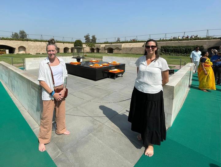 Two travellers stand beside a marble memorial platform adorned with orange flower trays in a manicured garden.