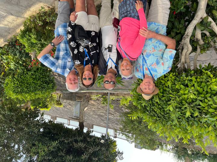 Five travelers wearing tour lanyards posing in a lush garden area with trimmed bushes and a stone wall behind.
