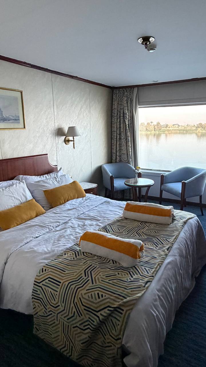 Cozy river-cruise cabin with neatly arranged bed and chairs facing large window over the Nile