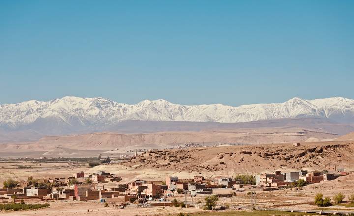 Adobe village at the foot of snow-capped Atlas Mountains under a bright blue sky.