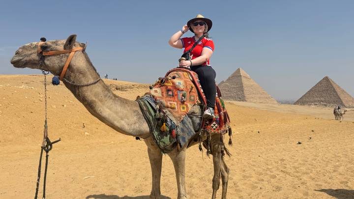 Traveler riding a camel in desert with Great Pyramids in the background.