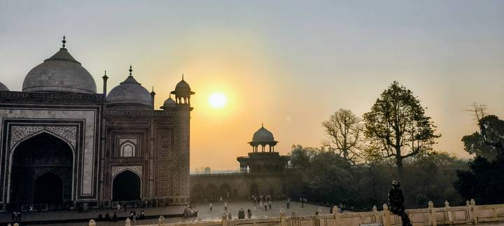Silhouetted Mughal pavilion beside the Taj Mahal complex at sunset with golden sun above.