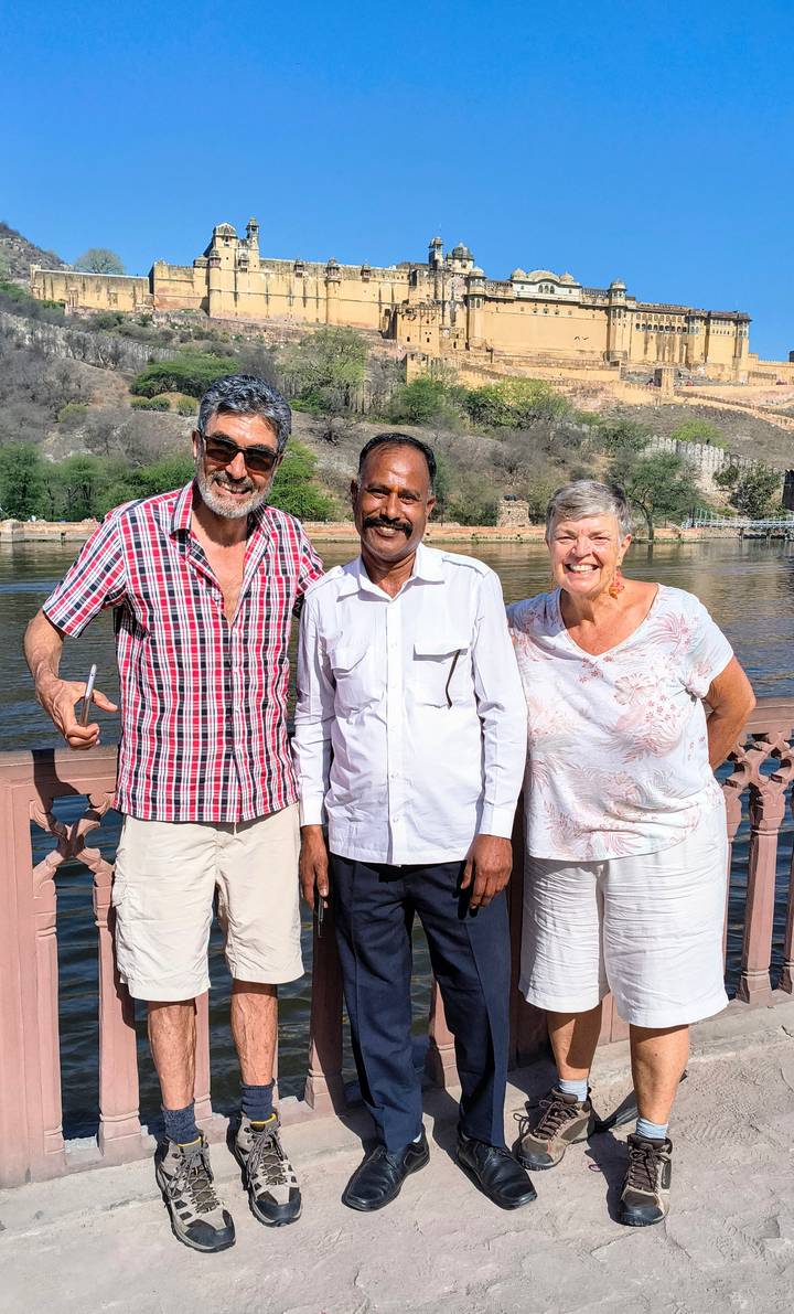 Two tourists smiling with their driver beside a riverside fort backdrop on a sunny day.