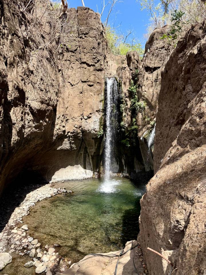 Narrow waterfall plunging into small pool within a rocky gorge lit by sunlight.