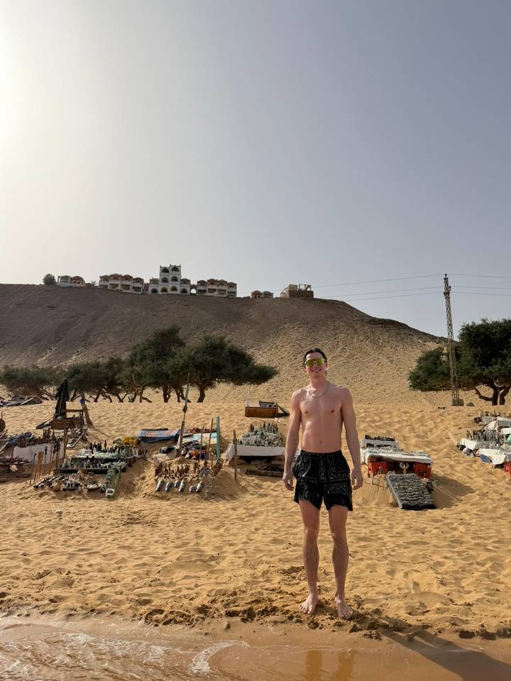 Shirtless traveler stands before a sandy hillside market on the edge of the desert.