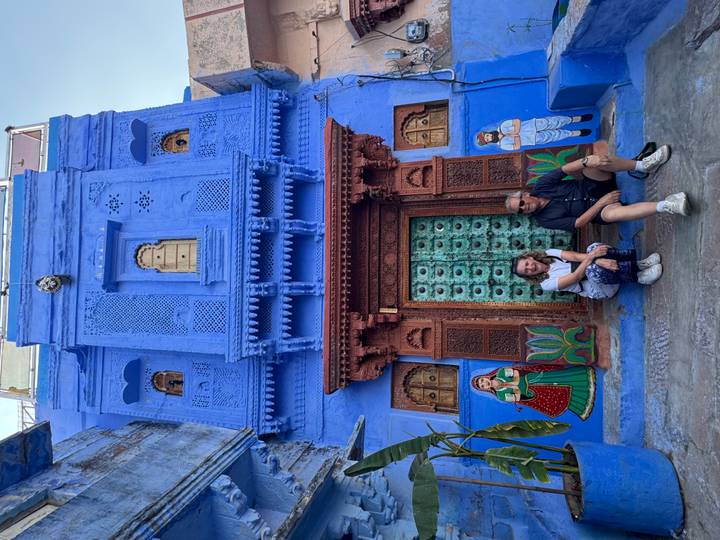 Couple seated in front of a vivid blue townhouse with ornate green door and folk murals in the Blue City.