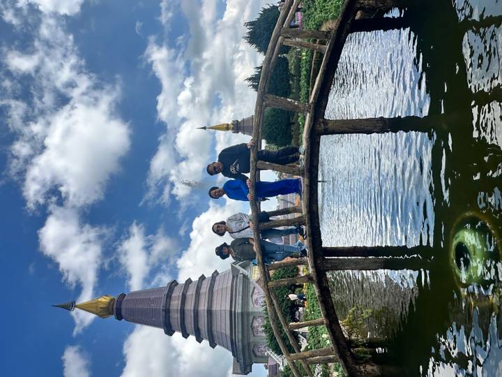 Four travellers standing on a small wooden bridge over a pond with twin pagodas behind.
