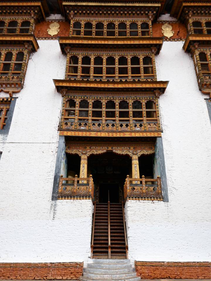 Facade of a Bhutanese monastery with richly carved and painted wooden balconies over a white wall.