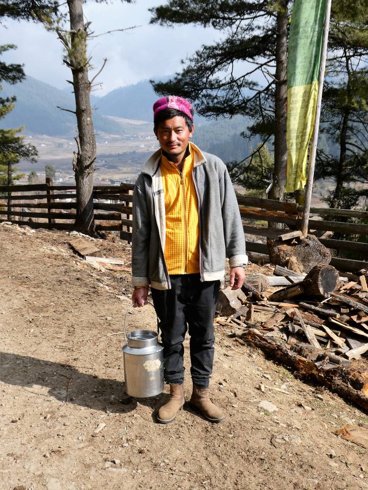 Bhutanese man carrying a metal bucket in a rural mountain setting with firewood piles behind.