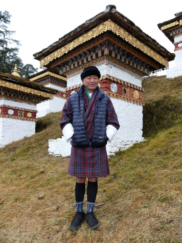 Bhutanese man in checked gho posing beside hillside chortens at a mountain pass.