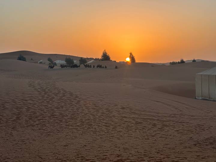 Orange sunset over Sahara sand dunes with a camel caravan and tents silhouetted on the horizon.
