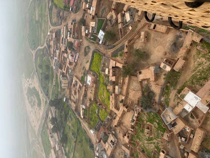 Hazy aerial view of a rural Moroccan village with green crop patches and winding dirt roads.