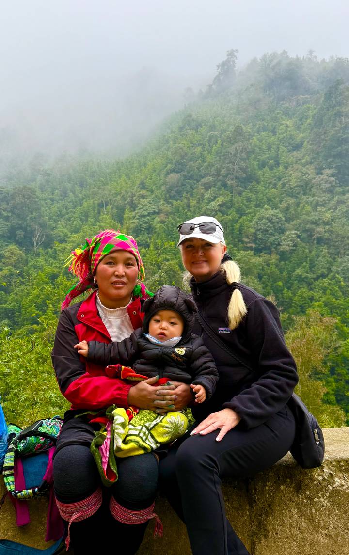 Two women and a baby in colorful ethnic attire stand against a lush mountainous backdrop in Sapa.