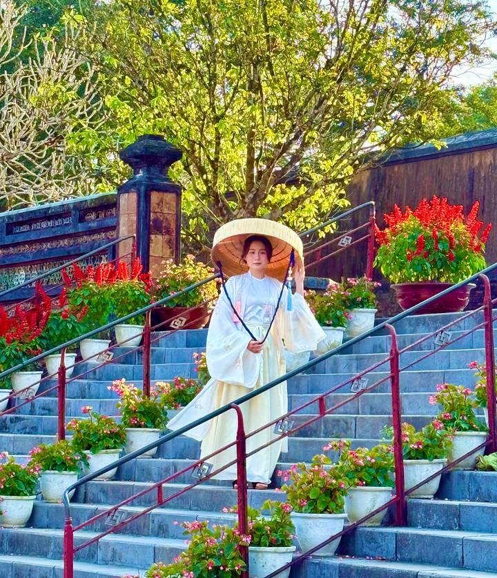 Woman in traditional white ao dai and conical hat stands gracefully on stone steps amid red flowers in Hue.
