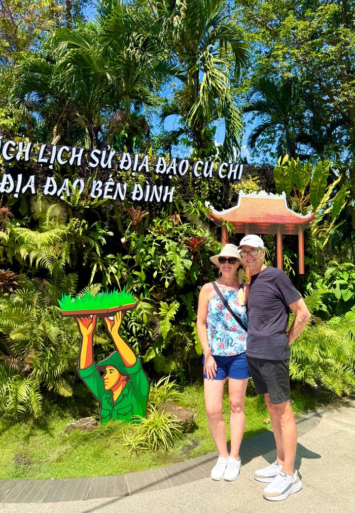 Couple poses below a Cu Chi Tunnels sign amid dense tropical foliage.