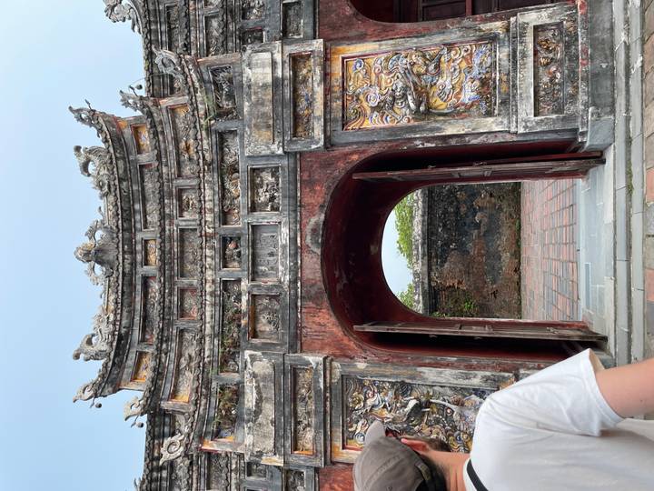 Weathered ornate stone gate with open doors; partial traveler visible at edge of frame.