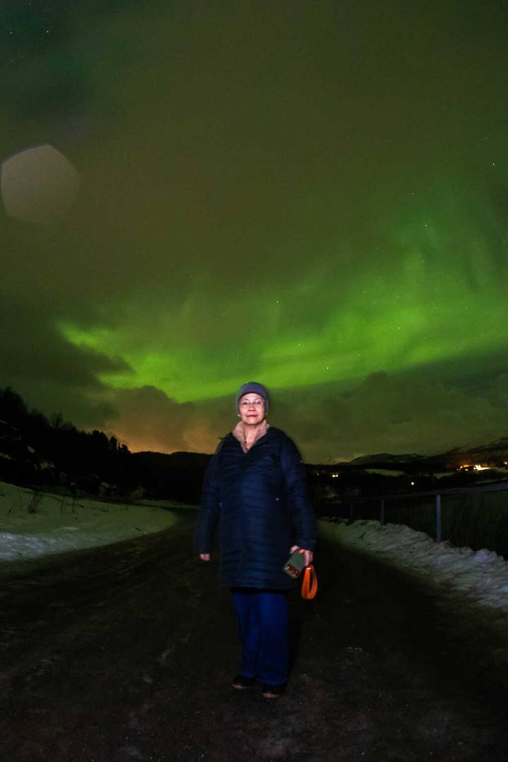 Portrait of a traveler standing under vivid green Northern Lights with snowy hills in Tromsø.
