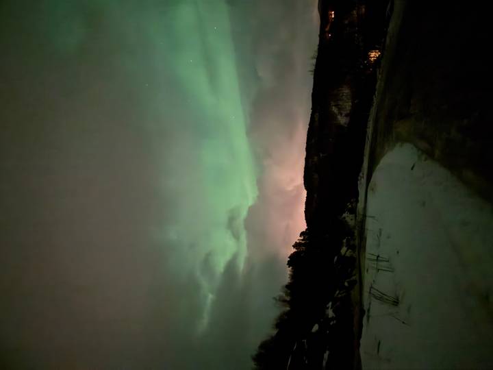 Green aurora borealis sweeping across a cloudy night sky above snowy terrain.