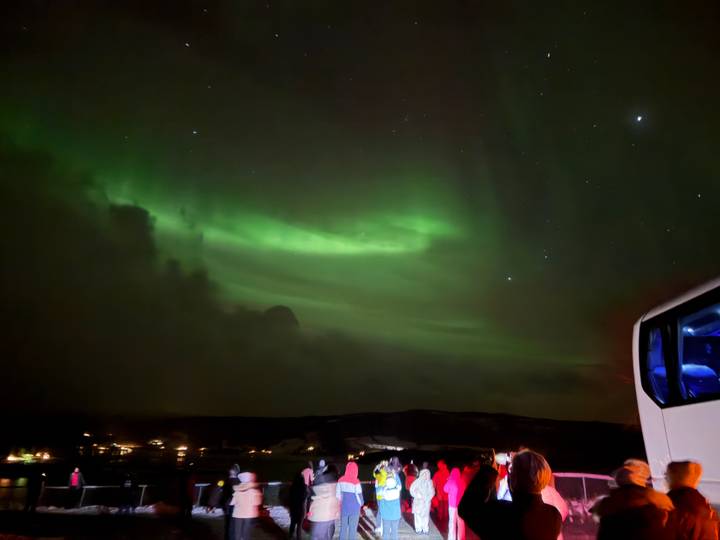 Green aurora lights dance across a star-filled night sky above snowy hills, with the window of a tour bus visible on the right.