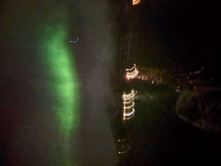 Very blurry image of green aurora and streaked lights above dark water at night.