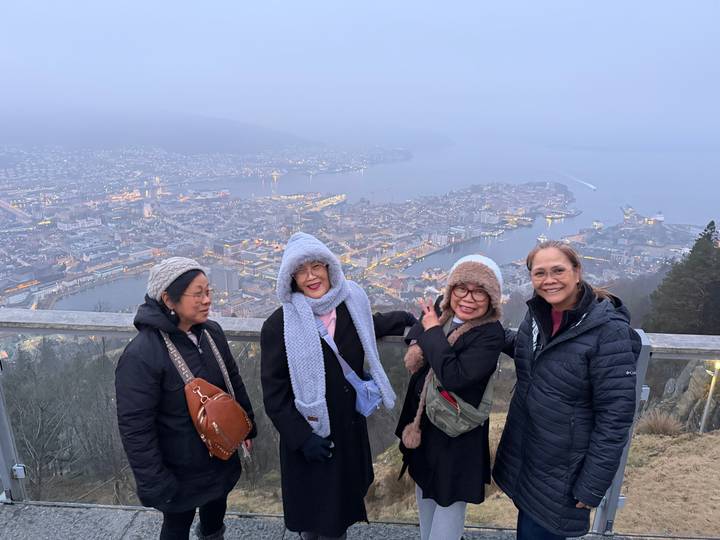 Four travellers pose at a mountain lookout rail above a misty coastal city and harbour.