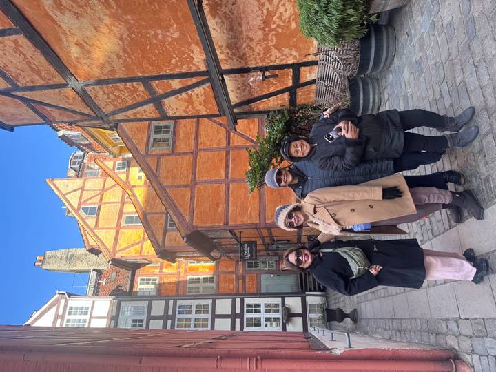 Four smiling women stand in a narrow cobblestone alley framed by bright orange half-timbered houses.