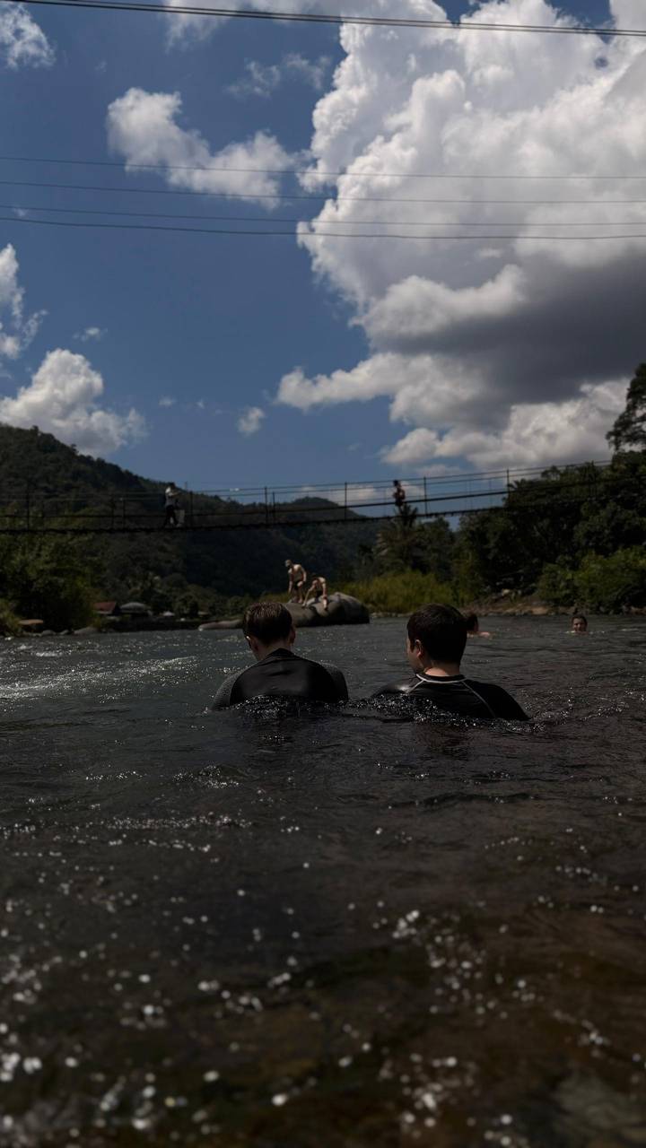 Travellers cool off in a jungle river below a hanging suspension bridge on a bright day.